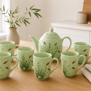 May include: A light green ceramic tea set consisting of a teapot and five matching mugs. Each piece features a hand-painted design of dark green grass-like stalks and small red fish. The teapot has a lid and a curved handle and spout.