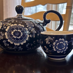 May include: A dark blue ceramic tea set with white floral patterns. The set comprises a teapot with a lid and two matching teacups. The tea set is displayed on a dark wooden surface.