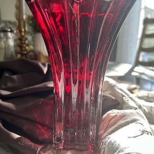 May include: A vibrant, ruby-red glass vase with a flared, scalloped rim. The vase features vertical ridges, creating a textured appearance. The vase is placed on a patterned surface.