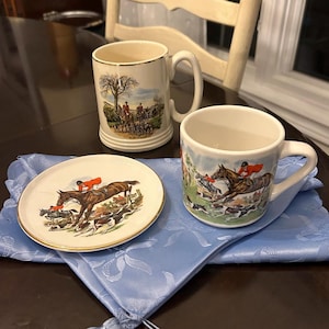 May include: A collection of vintage ceramic mugs and a small plate depicting hunting scenes. The mugs and plate showcase riders on horseback, hounds, and a fox hunt. The plate has a gold rim and rests on a light blue cloth.