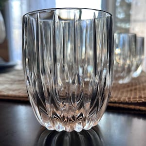 May include: Clear, textured glass tumblers with vertical grooves. The glasses are arranged on a brown woven placemat, with a blurred background. The glasses are on a dark wooden table.
