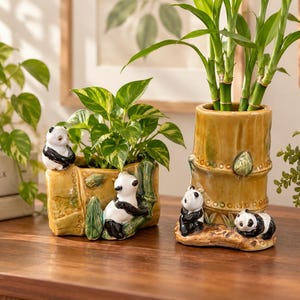 Peut inclure: Deux cache-pots en céramique en forme de tiges de bambou, ornés de petites figurines de pandas. Le pot plus court et large contient une plante verte, tandis que le pot cylindrique plus haut accueille des tiges de bambou. Finition jaune-brun.