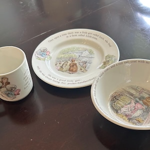 May include: A white ceramic mug, plate, and bowl set featuring illustrations of Beatrix Potter characters. The plate and bowl display colorful scenes with printed text from the stories, while the mug features a matching character graphic.