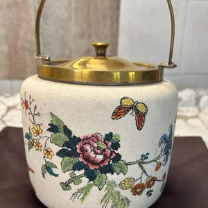 Peut inclure: Un pot en céramique crème avec un couvercle et une poignée en laiton. Le pot est décoré de motifs floraux et de papillons roses, verts, jaunes et oranges. Le couvercle est rond et possède un petit bouton en laiton.