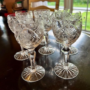 May include: A set of clear crystal wine glasses featuring intricate cut glass patterns and etched floral designs on the bowls. Each glass has a faceted stem and a circular base with a radiating starburst pattern, reflecting light through the glass.