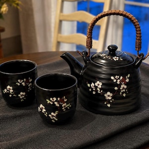 May include: A black ceramic tea set comprising a teapot and two matching cups. The teapot features a woven brown handle and a lid, both decorated with white cherry blossom patterns. The cups also display white cherry blossom designs.