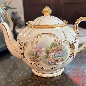 May include: A cream-coloured porcelain teapot featuring a gold-trimmed lid and handle. The body is decorated with a colourful landscape painting of a cottage surrounded by flowers, framed by ornate gold scrollwork accents on a fluted surface.