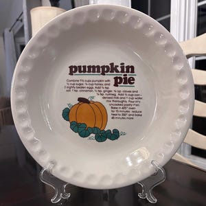 May include: A white ceramic pie plate with a decorative, scalloped edge. The plate features a graphic of a pumpkin and the words "pumpkin pie" in brown lettering. Recipe instructions are also printed on the plate.