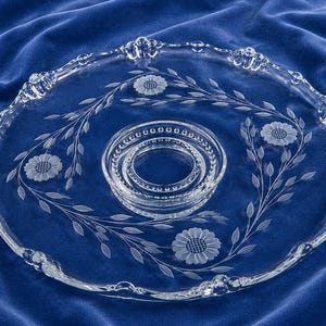 May include: A clear glass serving platter with a floral and leaf design. The platter has a scalloped edge and a raised centre section. The floral design includes etched sunflowers and delicate leaf patterns. The platter is approximately 30 cm in diameter.