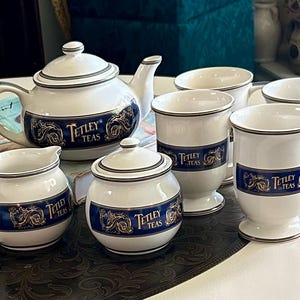 May include: A white ceramic tea set comprising a teapot, a creamer, a sugar bowl with a lid, and four mugs. Each item features a dark blue band with gold scrollwork and the text Tetley Teas in a gold font, finished with a silver-coloured trim.
