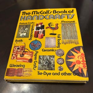 May include: A vintage book titled "The McCall's Book of Handcrafts" with a bright yellow cover. The cover features illustrations of various crafts, including candlemaking, batik, weaving, and macramé. The book is a guide to different crafting techniques.