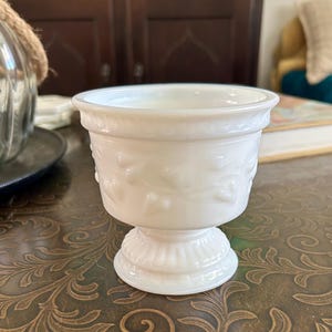 May include: A white milk glass pedestal bowl featuring a raised heart pattern around the exterior body, a flared rim with a decorative border, and a ribbed base. The vessel has a smooth, opaque finish and a classic footed design.