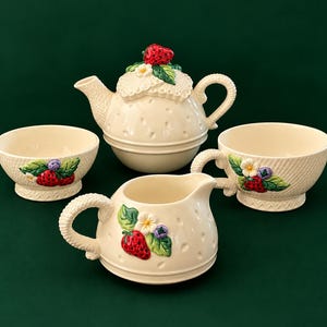 May include: A cream-coloured ceramic tea set featuring a teapot with a strawberry-shaped lid, a creamer, and two bowls. Each piece is decorated with raised, hand-painted strawberries, green leaves, white flowers, and small blue berries in relief.
