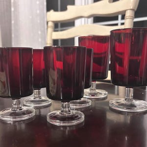 May include: A set of six ruby red stemmed glasses. The glasses have a cylindrical shape with vertical ridges and sit on clear, circular bases. The stem is clear and connects the base to the red glass. The glasses are arranged on a dark surface.