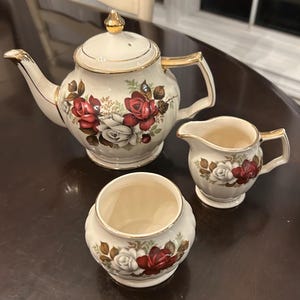 Peut inclure: Un service à thé en céramique de couleur crème comprenant une théière avec couvercle, un pot à lait et un sucrier. Chaque pièce présente un motif floral de roses rouges et blanches avec des feuilles vertes et un liseré doré sur les bords.