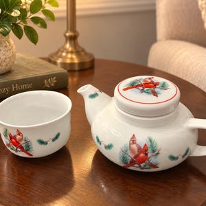 May include: A white ceramic teapot and matching teacup set featuring a festive cardinal bird and green pine branch pattern. The teapot includes a round lid with a red rim, while both pieces display detailed illustrations of red cardinals.