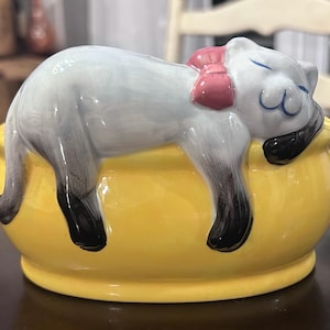 May include: A ceramic figurine of a sleeping cat with a pink bow, resting on a yellow bowl. The cat has blue eyes and black paws. The bowl is a bright yellow color.