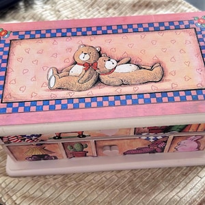 May include: A pink wooden jewelry box with a rectangular lid featuring a painted scene of two teddy bears. The lid has a blue and white checkered border and floral accents. The box has several drawers with illustrated panels.