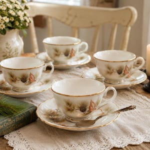 May include: Four white teacups and saucers with gold rims and a pinecone and leaf design. A vintage spoon rests on one saucer. A lit candle and a vase of flowers are in the background.
