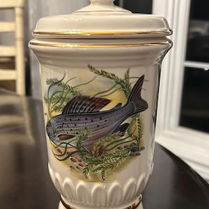 May include: A cream-coloured ceramic jar with a lid, featuring a detailed illustration of a fish. The jar has gold trim and a decorative base. The fish is depicted in shades of grey, brown, and blue, surrounded by aquatic plants.