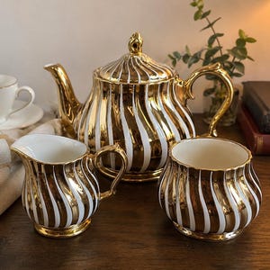 May include: A gold and white striped tea set, comprising a teapot, creamer, and sugar bowl. The set has a glossy finish and is on a wooden surface. A white teacup and saucer are in the background.