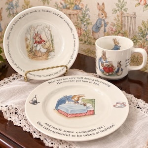 May include: A ceramic dinnerware set featuring Peter Rabbit illustrations. The bowl reads, Then old Mrs. Rabbit took a basket and her umbrella, and went through the wood to the baker's. The plate reads, Peter was not very well during the evening. His mother put him to bed. One table-spoonful to be taken at bed-time.