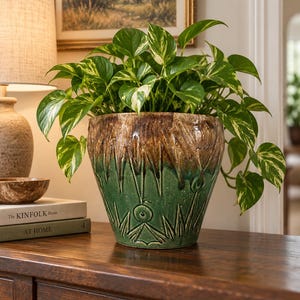 May include: A ceramic plant pot with a tapered shape, featuring a two-tone glaze of dark brown at the top and vibrant green at the bottom. The surface has a textured, carved pattern of vertical spikes and circular motifs, containing a leafy pothos plant.