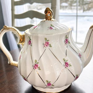 Peut inclure: Théière en porcelaine blanche avec des détails dorés sur la poignée, le bec et le couvercle. Elle est ornée de motifs de roses roses et de feuilles vertes, ainsi que de lignes dorées en treillis. La théière est posée sur une surface marron foncé.