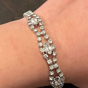 May include: A silver bracelet adorned with clear, sparkling rhinestones. The bracelet showcases a pattern of round and marquise-cut stones in a symmetrical design. The bracelet is displayed on a wrist.