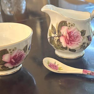 May include: A white porcelain cream jug, a matching sugar bowl, and a small spoon, all decorated with pink rose blossoms, green leaves, and gold-toned trim. The cream jug features a curved handle and a pouring spout, while the spoon handle has a floral pattern.