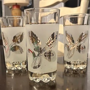 May include: Three vintage frosted glass shot glasses with gold rims and wheat stalk designs. The wheat stalks are in shades of gold, green, and silver. The glasses have a decorative base.