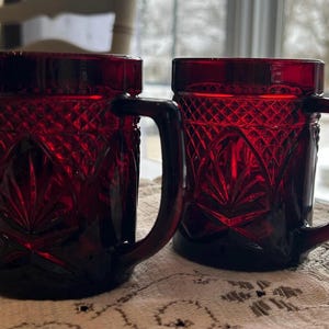 Peut inclure: Deux chopes en verre rouge rubis avec un motif de losanges et d'éventails. Chaque chope a une anse et une base épaisse. Les chopes sont posées sur une nappe en dentelle blanche. Les chopes sont d'un rouge profond.