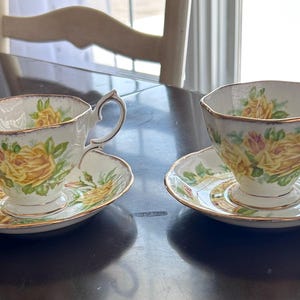 May include: Two vintage teacups and saucers. The cups and saucers are white with a yellow rose pattern and gold trim. The teacups have a unique handle design. The set is placed on a dark surface.