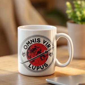 May include: White ceramic mug with a handle, featuring a design with the words "OMNIS VIR LUPUS" around a red circle with a sword. The mug is on a wooden surface, next to a laptop and a small potted plant.