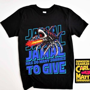Może przedstawiać: Czarny t-shirt z grafiką hybrydy rekina-pająka ziejącej ogniem, napisem "JAMAL" w kolorze niebieskim i frazą "HAS SO MANY SORRIES TO GIVE". Widoczna okładka książki "Dungeon Crawler".