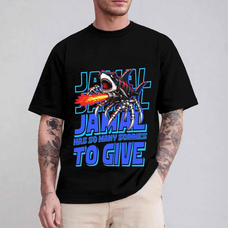 Może przedstawiać: Czarny t-shirt z grafiką hybrydy rekina-pająka, kt&oacute;ra zionie ogniem. Tekst "JAMAL HAS SO MANY SORRIES TO GIVE" jest niebieski z białym obrysem.
