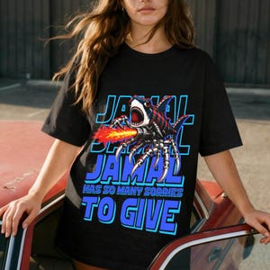 Może przedstawiać: Czarny t-shirt z graficznym wzorem hybrydy rekina-pająka z miotaczem ognia. Tekst "JAMAL HAS SO MANY SORRIES TO GIVE" jest w kolorze niebieskim i białym.