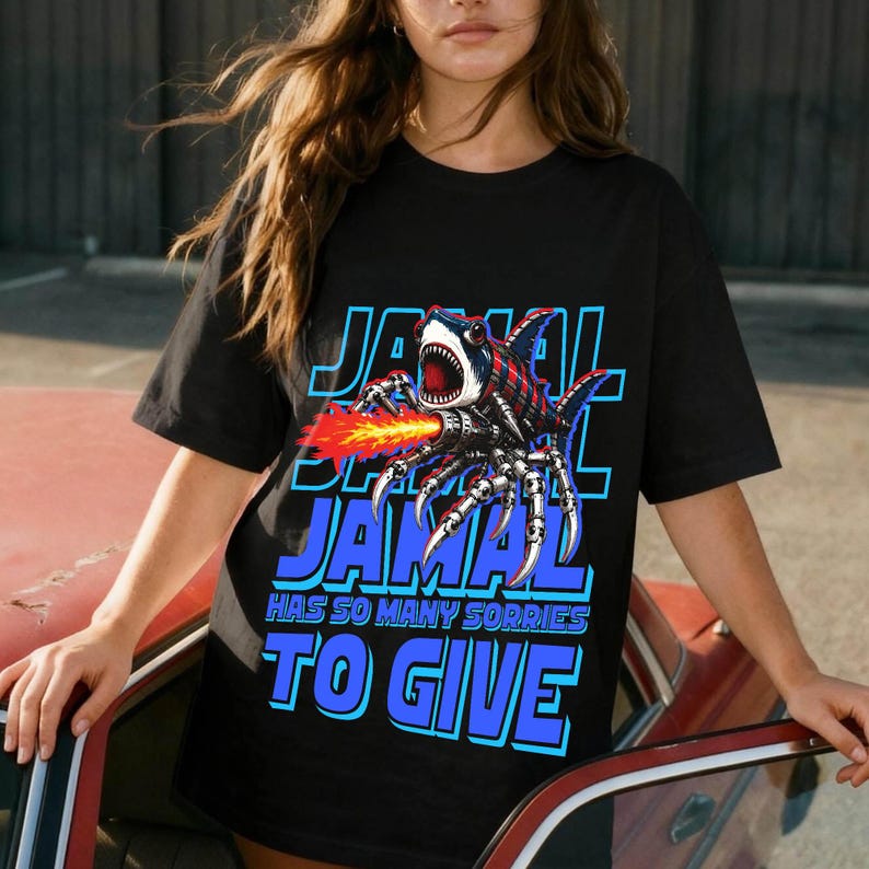 Może przedstawiać: Czarny t-shirt z graficznym wzorem hybrydy rekina-pająka z miotaczem ognia. Tekst "JAMAL HAS SO MANY SORRIES TO GIVE" jest w kolorze niebieskim i białym.