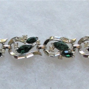 May include: A silver bracelet with a delicate design featuring green and clear rhinestones. The bracelet is made up of repeating sections of interlocking leaves and swirls.