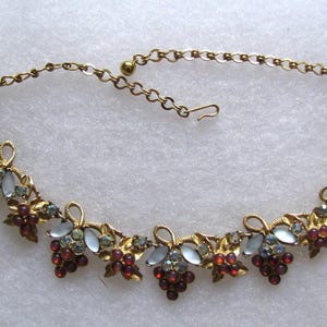 May include: A gold necklace with a grapevine design featuring red and blue rhinestones. The necklace has a delicate chain and a clasp closure.