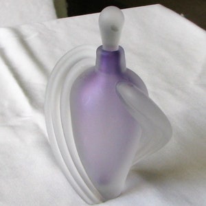 May include: A frosted glass perfume bottle with a purple hue and a unique, abstract design. The bottle has a frosted glass stopper and a curved, flowing shape.