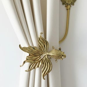 May include: A decorative gold-coloured curtain tieback shaped like a goldfish. The tieback is attached to a wall bracket with a floral design. The curtain is a light cream colour.