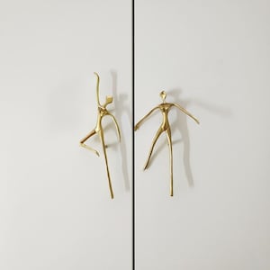 Puede incluir: Dos tiradores dorados para armario con forma de figuras humanas estilizadas. Una figura posa con un brazo levantado, la otra con los brazos extendidos. Los tiradores están montados en una puerta de armario blanca.