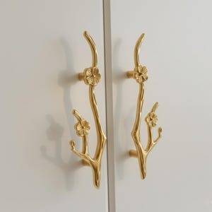 Op de afbeelding: Twee goudkleurige kastgrepen. Elke greep is gevormd als een tak met kleine bloemaccenten. De grepen zijn gemonteerd op een witte kastdeur en bieden een decoratief en functioneel element.