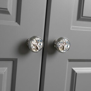 May include: Two cabinet knobs with a steampunk design. The knobs are silver-toned with a clear face revealing the intricate gears and mechanisms of a watch. The knobs are mounted on a gray cabinet door.