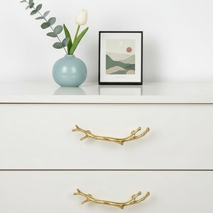 Op de afbeelding: Een witte dressoir met twee lades, elk met een goudkleurige takvormige handgreep. Een lichtblauwe vaas bevat eucalyptus en een witte tulp. Een ingelijste landschapskunstafdruk staat erop.