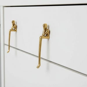 May include: Gold-coloured cabinet pulls shaped like a person sitting with legs extended. They are mounted on a white cabinet. The design is minimalist and artistic, adding a unique touch to furniture.