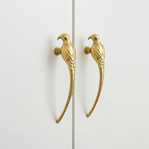 Puede incluir: Dos tiradores dorados para armario con forma de pájaro. Los tiradores están montados en una puerta de armario blanca. Los tiradores de pájaro tienen diseños detallados de plumas y picos, añadiendo un toque decorativo.