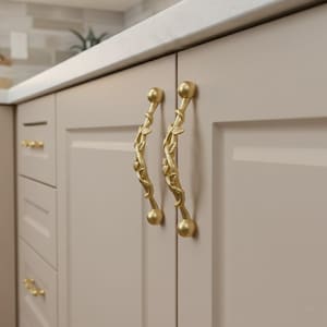 Könnte beinhalten: Zwei goldfarbene Schrankgriffe mit einem botanischen Design. Die Griffe sind gebogen mit einem Blatt- und Rankenmuster und haben an jedem Ende einen runden Knopf. Die Schranktüren sind hellbeige und die Arbeitsplatte ist weiß.