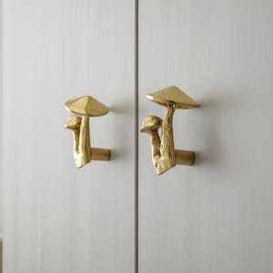 Könnte beinhalten: Zwei goldfarbene Pilz-Schrankgriffe an einer weißen Schranktür. Die Griffe haben ein strukturiertes, organisches Design mit einer großen Kappe und einem kleineren Pilz daneben. Die Schranktür hat ein vertikales Holzmaserungsmuster.